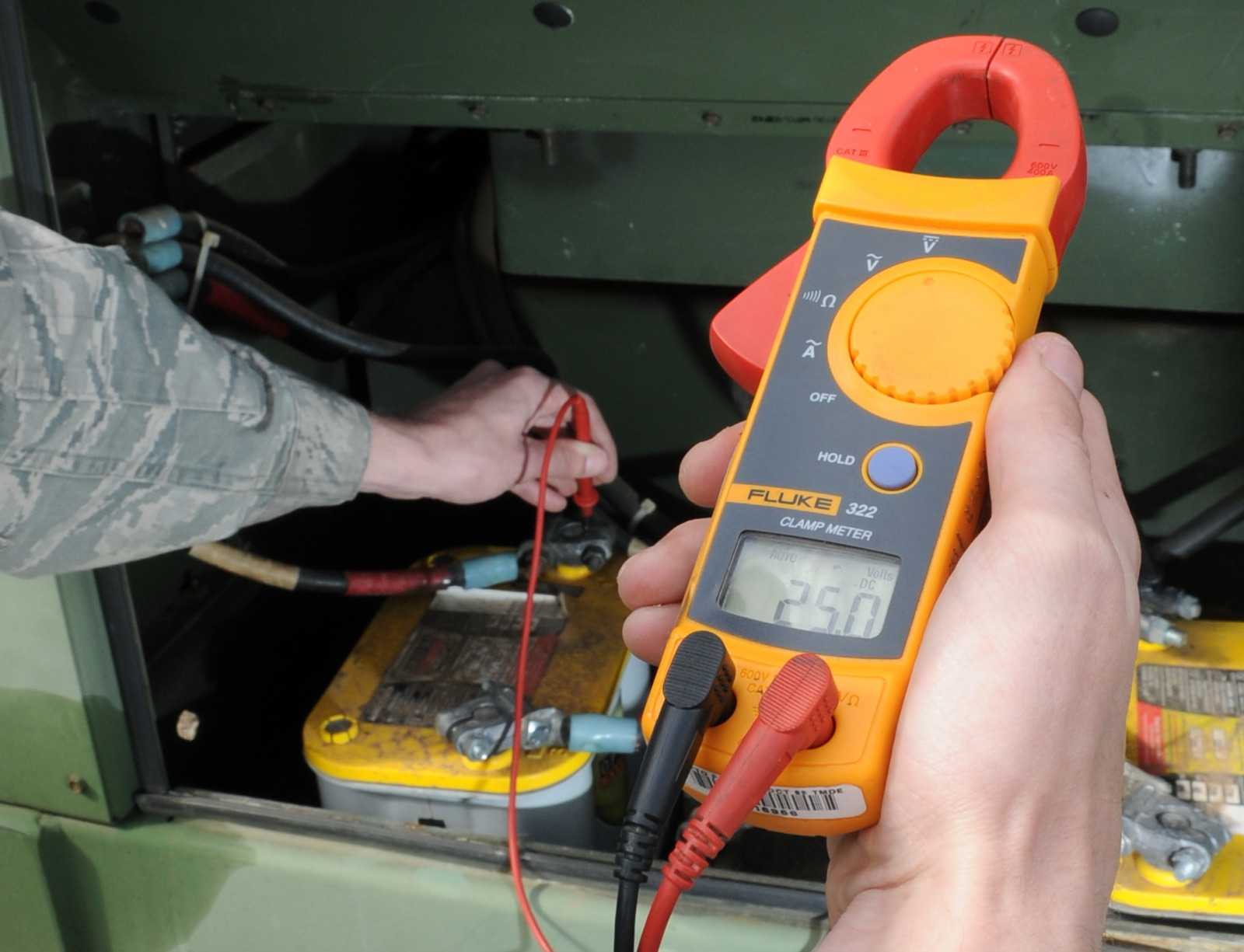 Hands holding a clamp meter around a cable on a battery bank, with test leads also connected to measure voltage