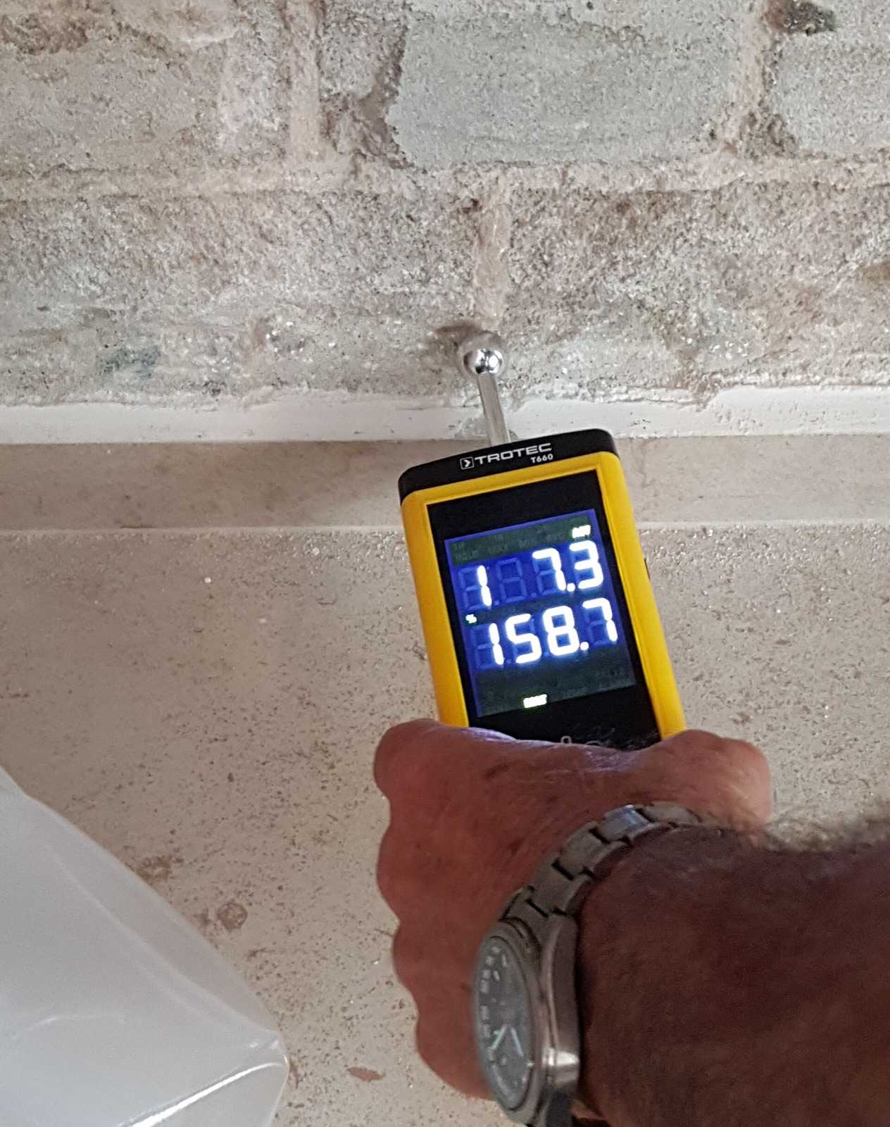 Hand pressing a pin-type moisture meter into a wall, the digital display showing a reading