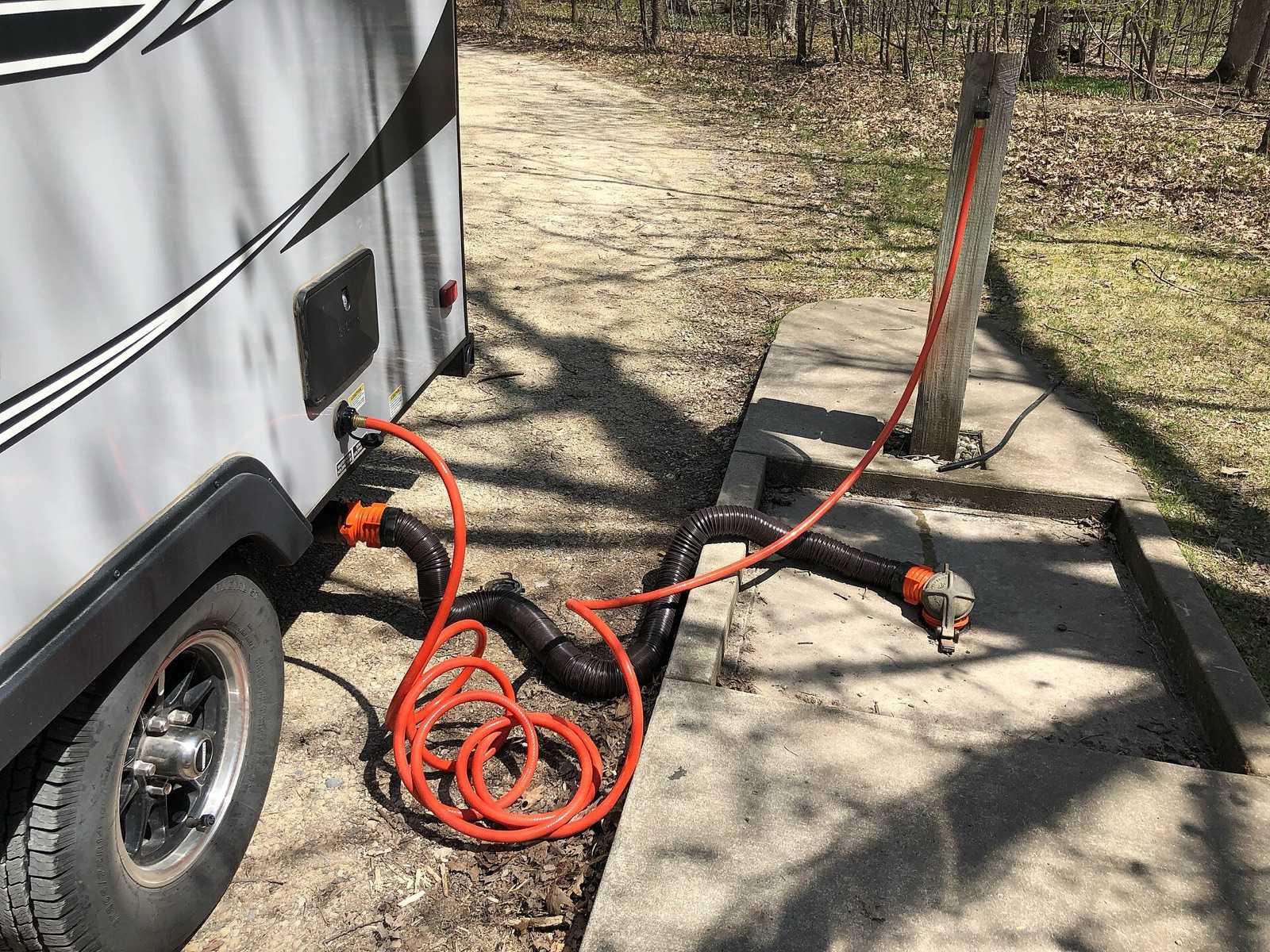 Travel trailer at a campground dump station with a black sewer hose running to the drain inlet and an orange non-potable hose connected to the trailer's tank rinser fitting