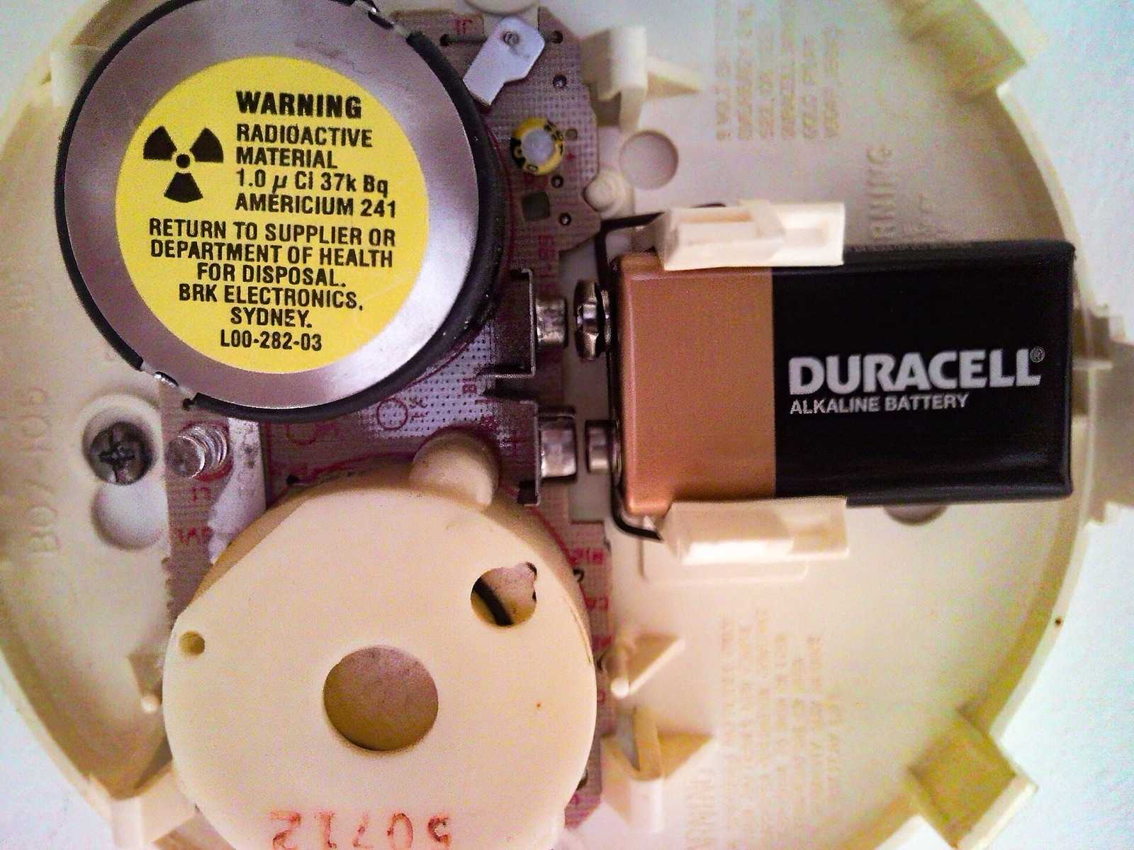 Inside of a smoke alarm with the cover off, showing the 9 V battery and the ionization chamber with its warning label