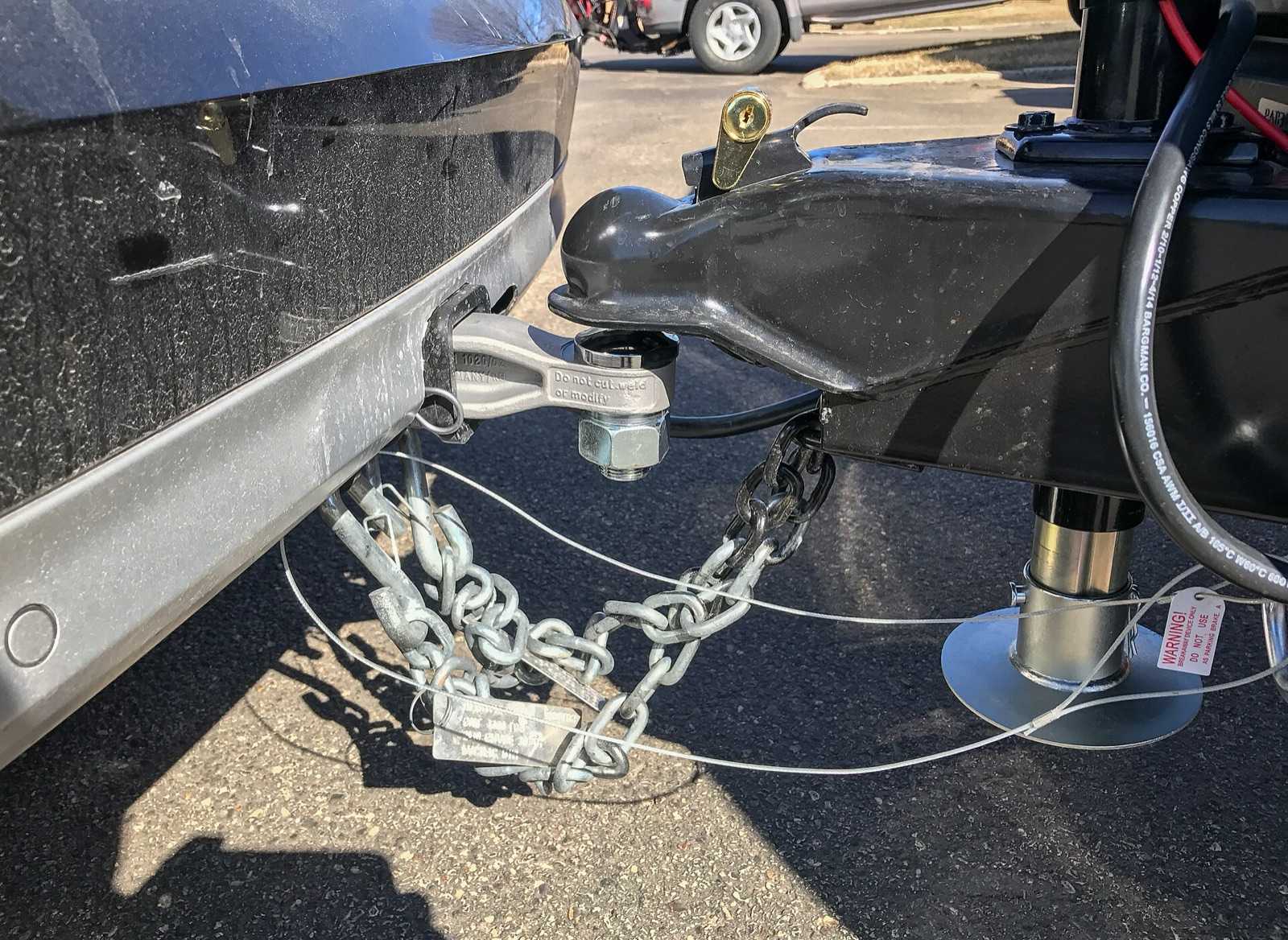 Trailer coupler latched on a hitch ball with a lock pin, safety chains crossed under the coupler, and a breakaway cable with a warning tag clipped separately to the truck