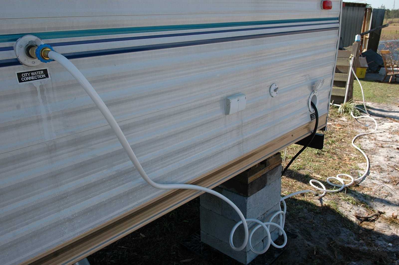 White drinking-water hose threaded onto a travel trailer's city water connection inlet, with the shore power cord entering the sidewall nearby