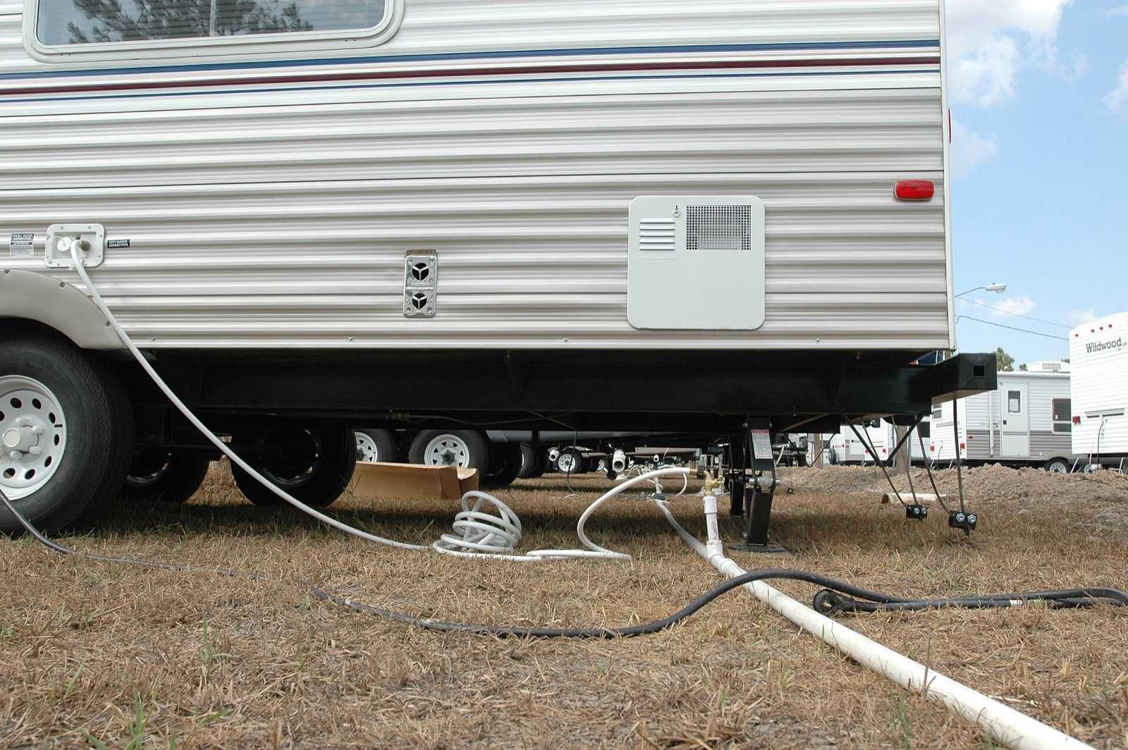 Low-angle view of a travel trailer's curb side showing the water heater access door with its vent grille, the chrome furnace intake and exhaust vent, and a hose connected to the city water inlet