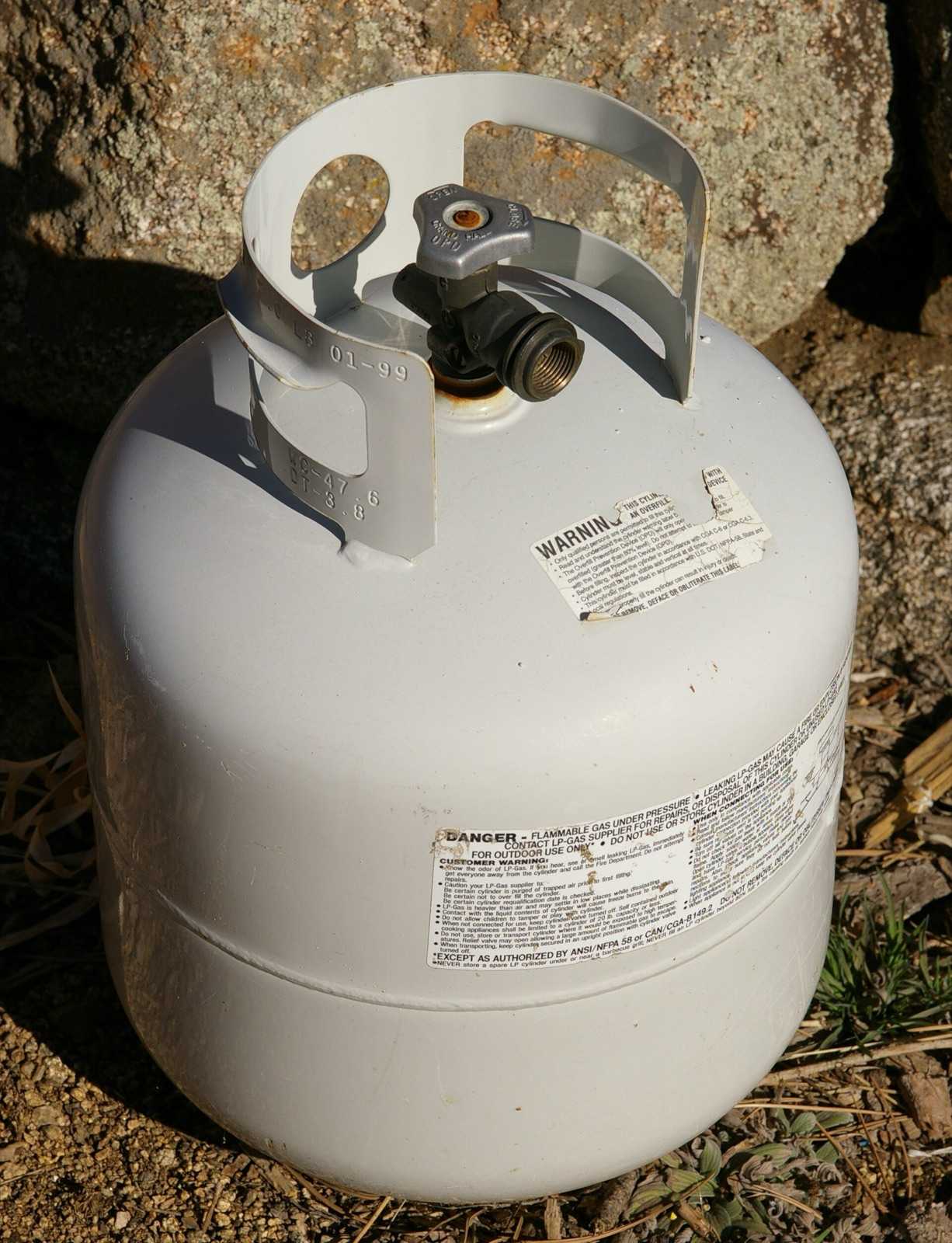 White 20 lb propane cylinder with an OPD valve and protective collar, sitting outdoors on rocks